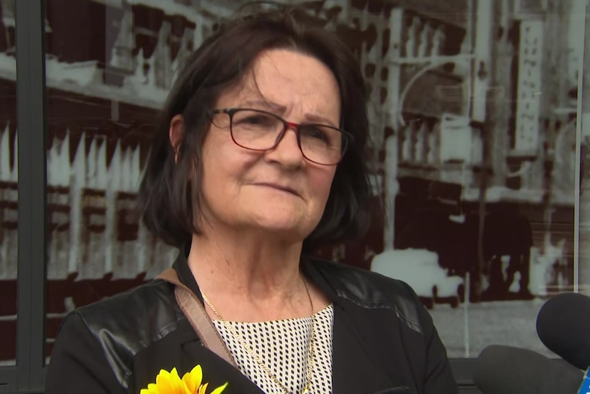 Woman wearing glasses with sunflower in jacket lapel standing outside glass building.