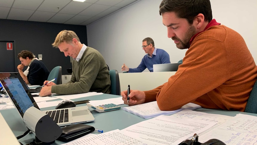 Four men sitting at a table writing down prices as they bid on wool.