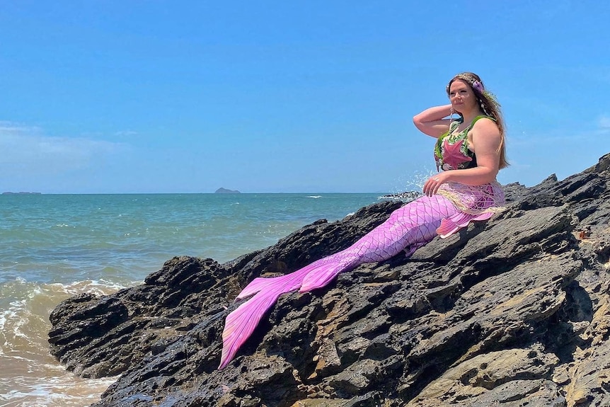 A woman with a pink mermaid tail sits on some rocks at the beach