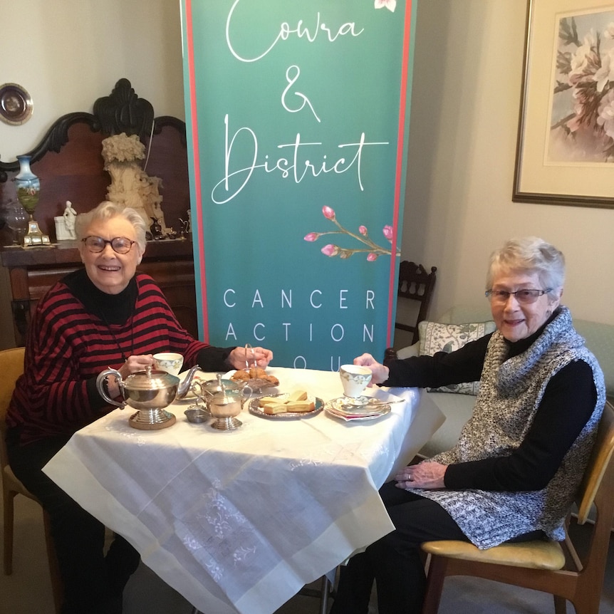 Two ladies sitting infront of a Cowra Cancer Action Group sign with tea and scones