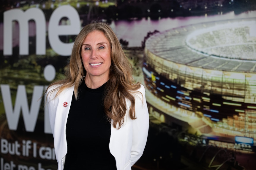 A lady with long light brown hair wears a white jacket and is pictured in front of a photo of Perth Stadium
