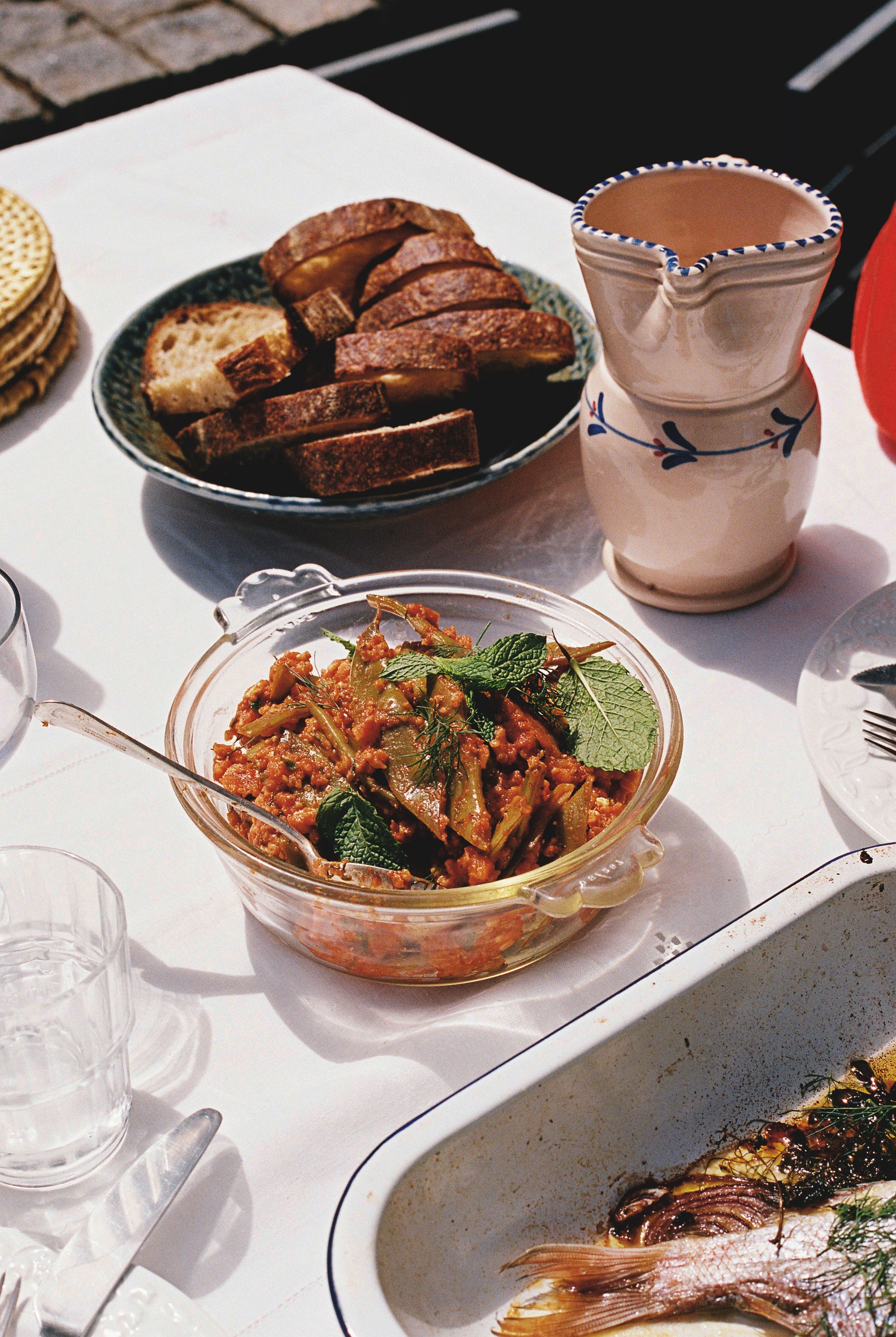 A glass bowl of green beans in red sauce with a grain mixed through, on a table with a white cloth and bread