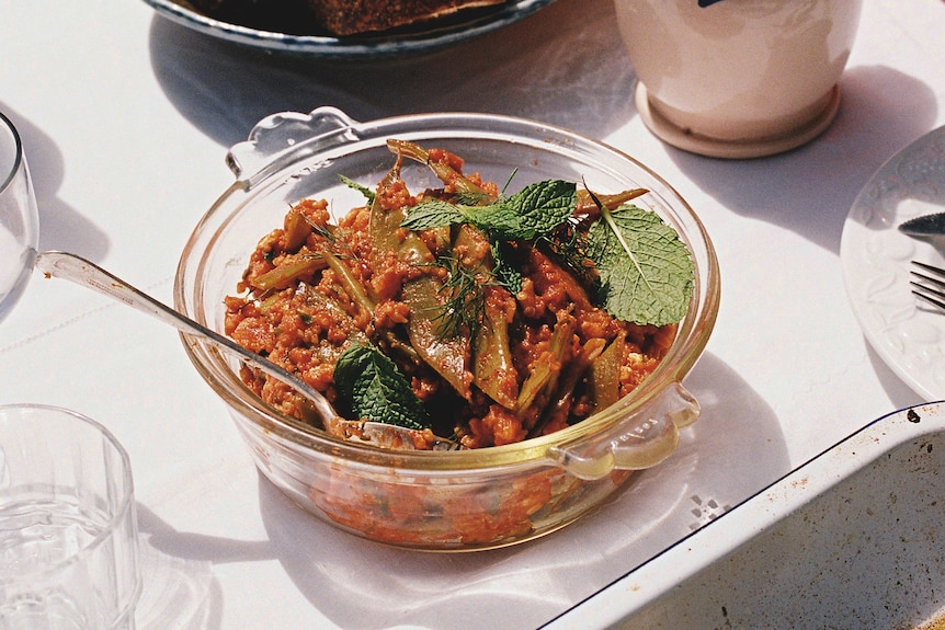 A glass bowl of green beans in red sauce with a grain mixed through, on a table with a white cloth and bread