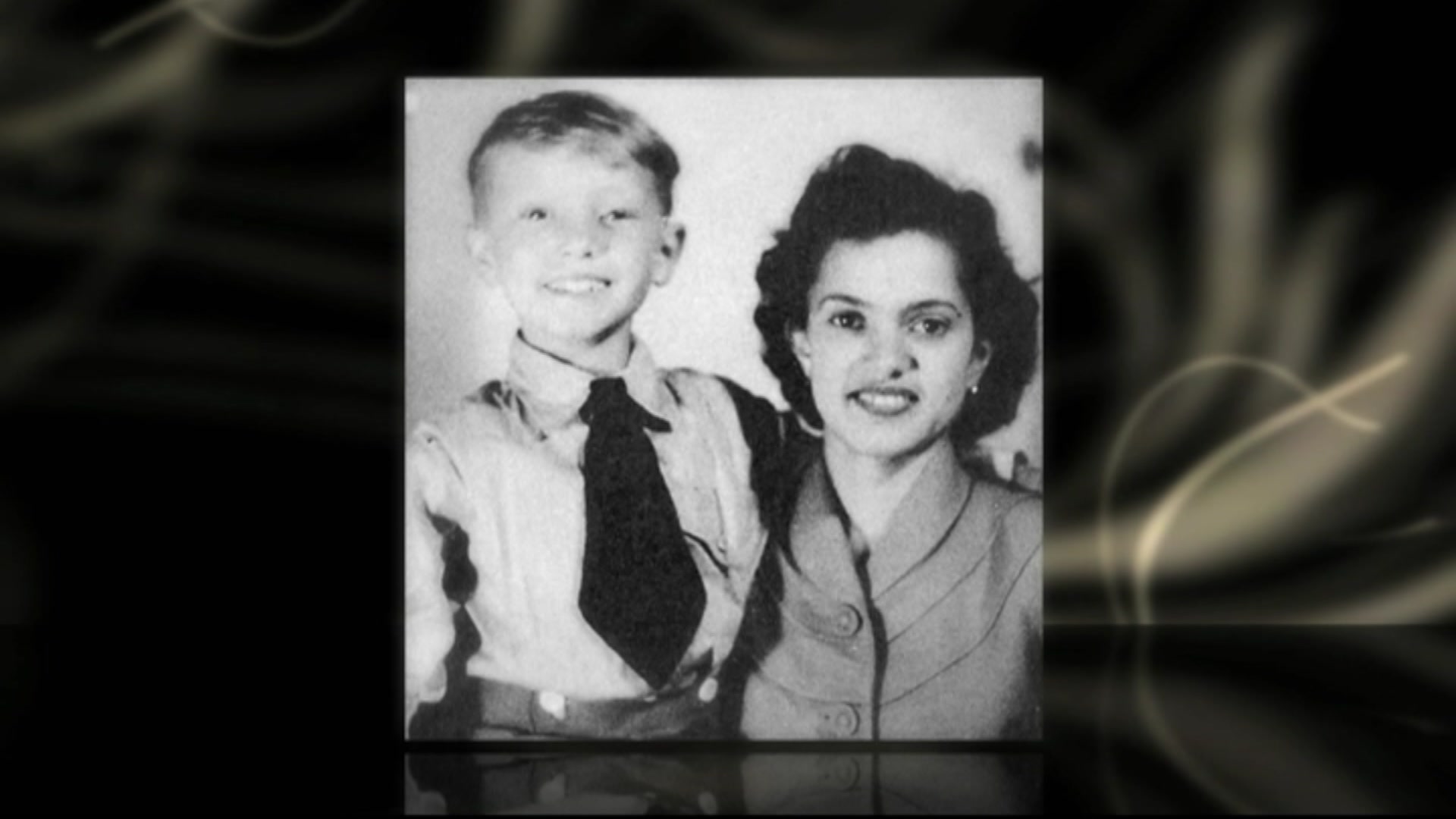 A black and white photo of a young boy in a collared shirt and tie standing with his arm around a woman in a dress.