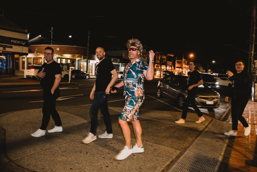 Five young men walk the street of Byron Bay, with four in all black with white shoes, and one with a vibrant shirt and shorts 