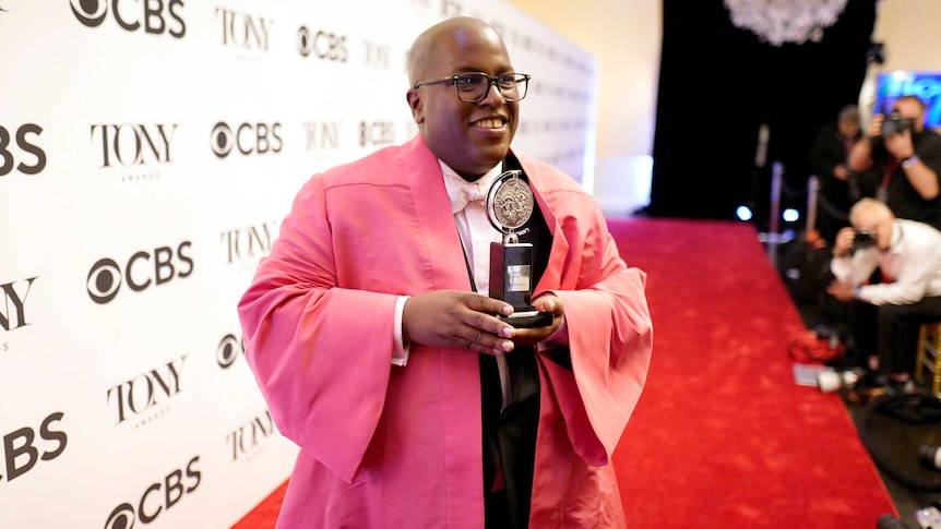 Michael R. Jackson in a tuxedo and pink overcoat stands on a red carpet and presents his Tony Award to a media scrum.