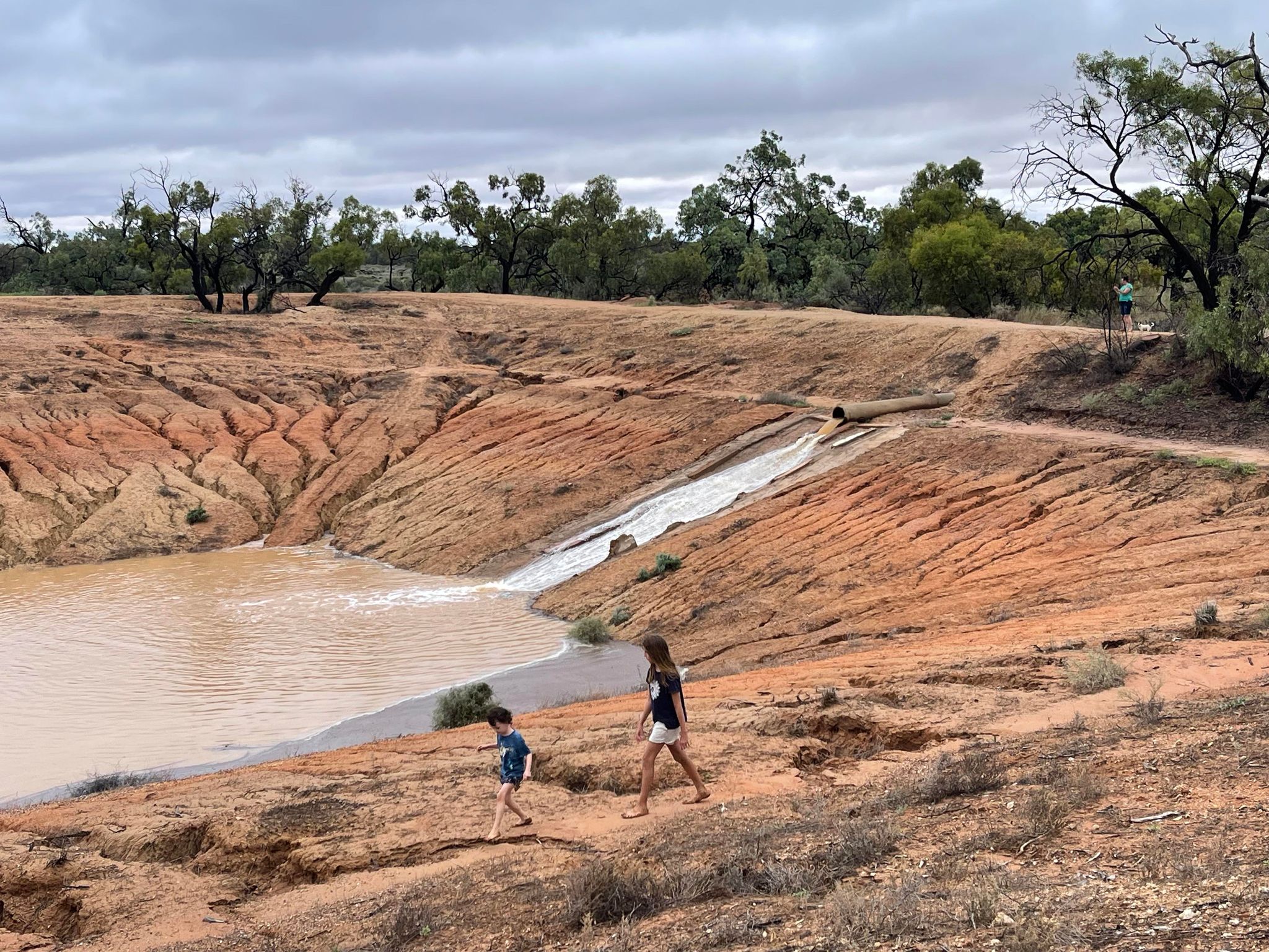 Two kids watch as water pours into a red-dirt dam in the outback.