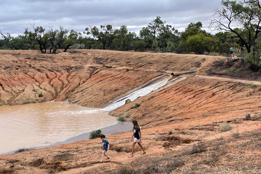 Two kids watch as water pours into a red-dirt dam in the outback.