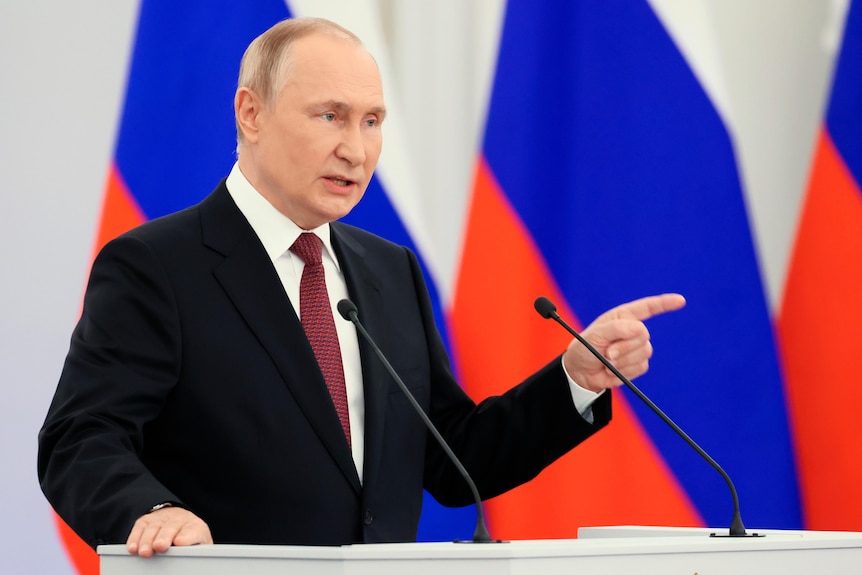 Vladimir Putin pointing as he speaks from podium.
