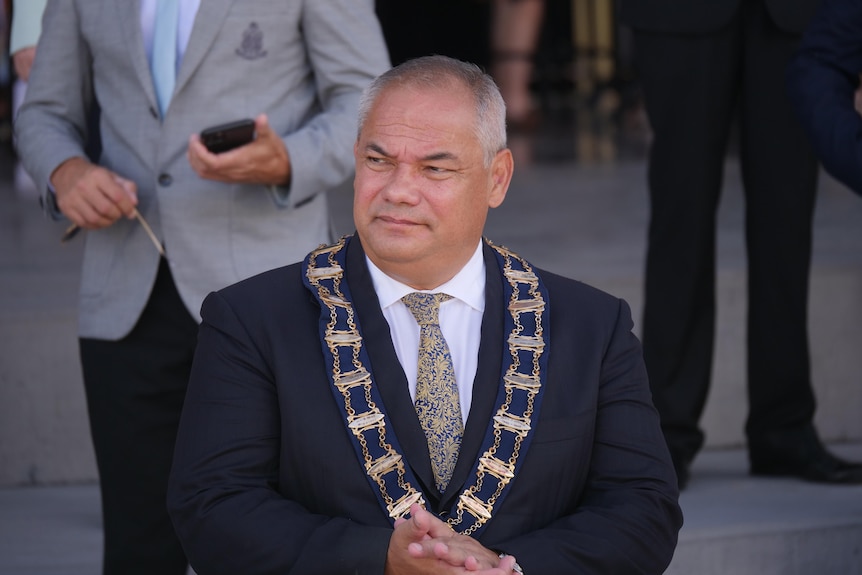 A bald man wearing a dark suit and mayoral livery sits down at a public event.