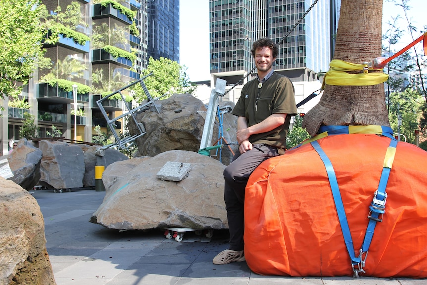 A man smiles as he sits on a big orange tarp with rocks in the background