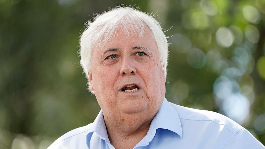 A close up of Clive Palmer wearing a blue business shirt in focus with green trees blurred in the background.