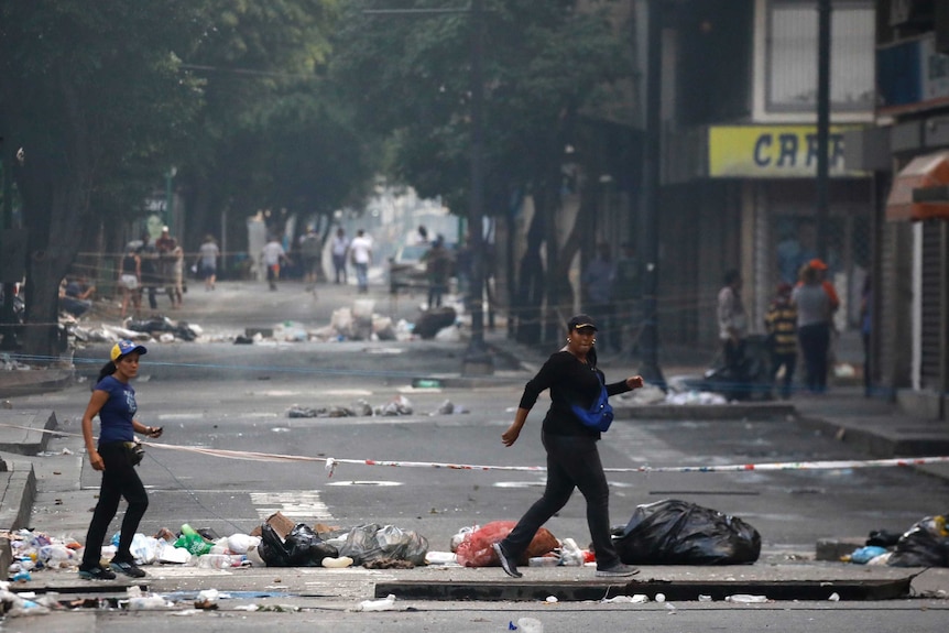 Two women walk across a street strewn with rubbish.