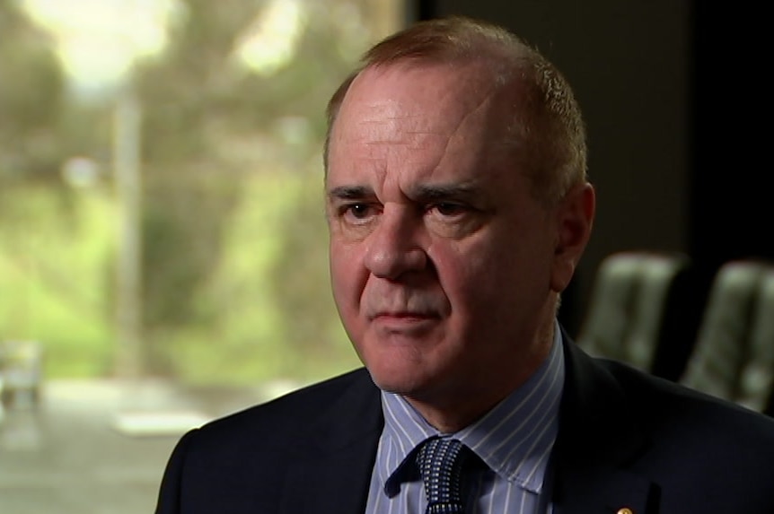 A man in a suit and jacket is interviewed while sitting on a chair.