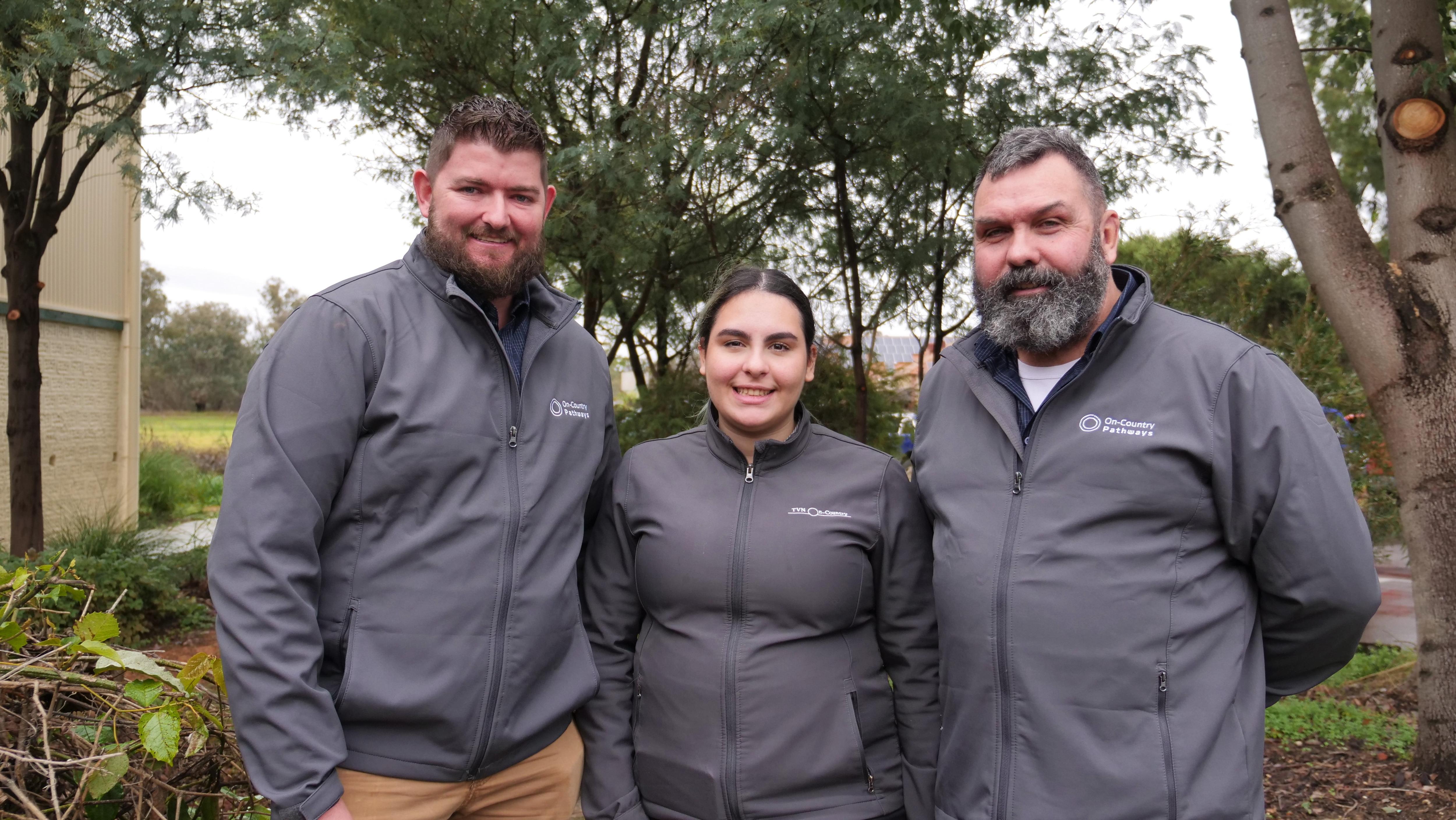 A young woman stands in between two men all in matching grey jackets outside in a garden.
