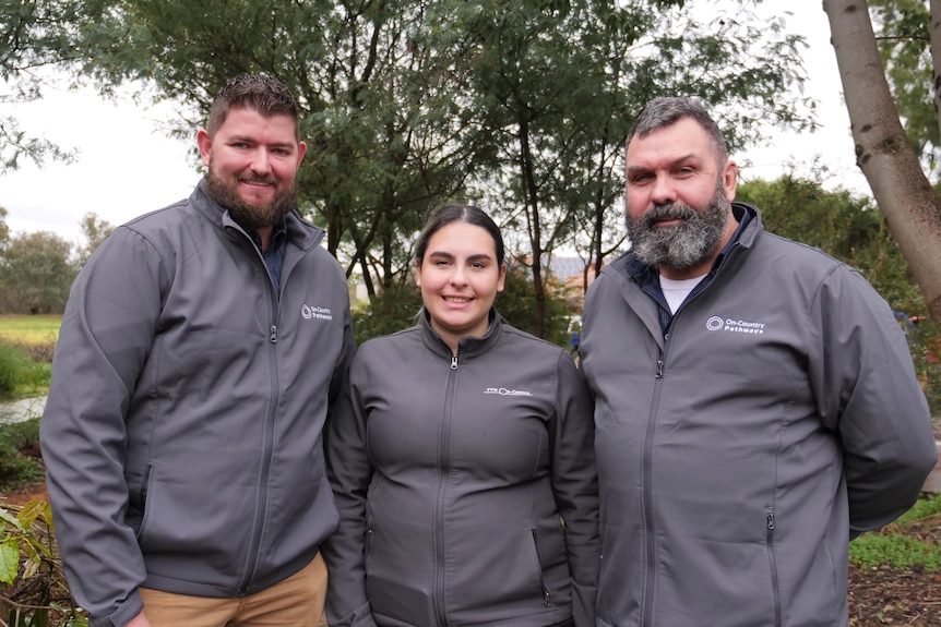 A young woman stands in between two men all in matching grey jackets outside in a garden.