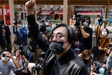 A masked man in a suit stands before a crowd of journalists and onlookers. He has raised his fist above his head