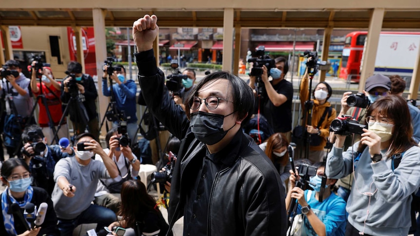 A masked man in a suit stands before a crowd of journalists and onlookers. He has raised his fist above his head