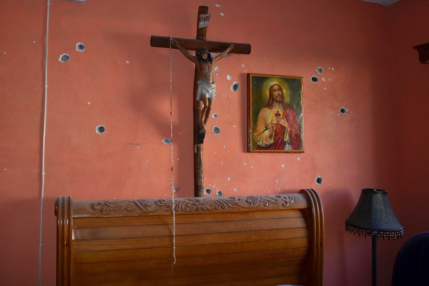 A religious cross and a painting hang on a wall of a home beside bullet holes.