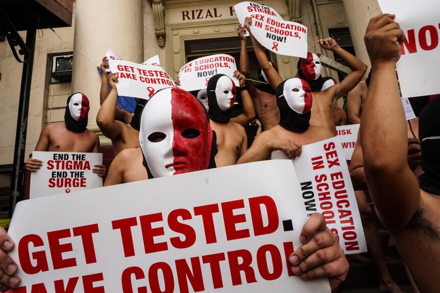 A group of shirtless men with faces painted red and white hold posters advocating for HIV testing and education. 