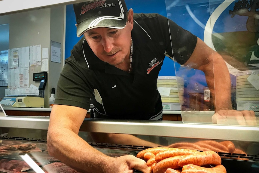 A man leans into the meat display cabinet at his butcher shop to place some sausages.