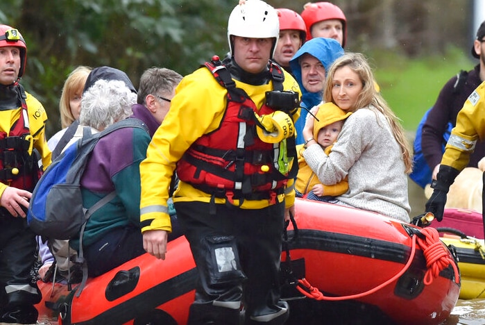 Two men, in yellow and red waterproof gear, guide an inflatable raft carrying rescued people through a flooded street in a town.