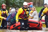 Two men, in yellow and red waterproof gear, guide an inflatable raft carrying rescued people through a flooded street in a town.