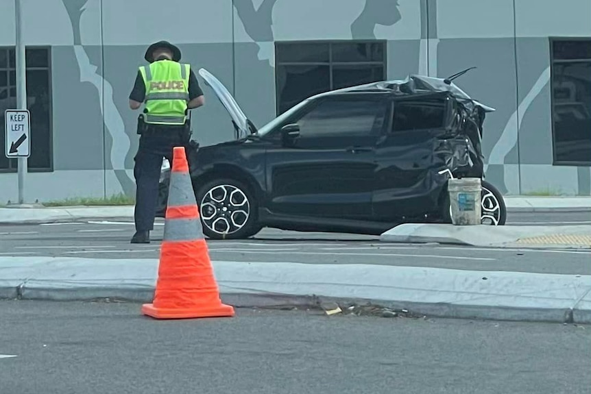 A police officer standing next to a black hatchback that has been hit from behind and badly damaged.