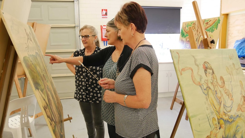 Three women stand looking at a Pixie O'Harris painting.