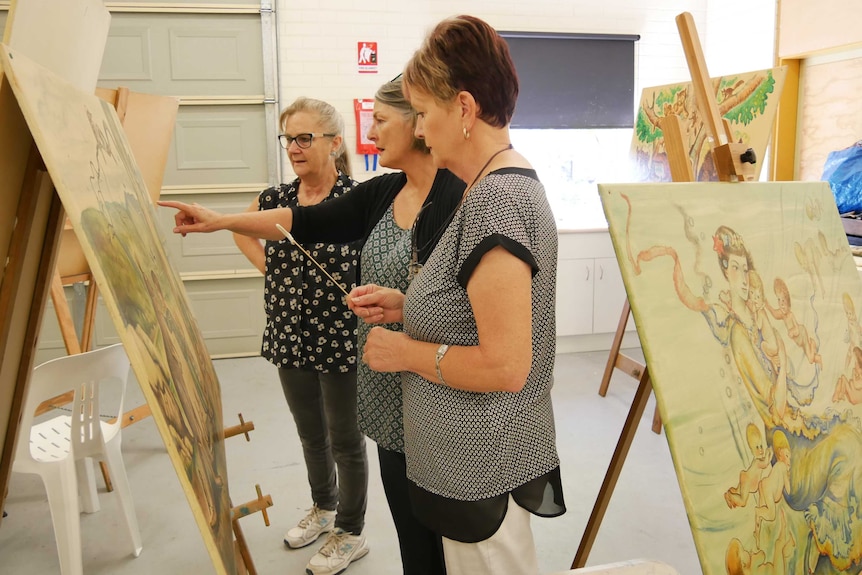 Three women stand looking at a Pixie O'Harris painting.