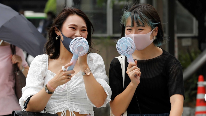 Two women wearing masks blow fans on their faces as they walk