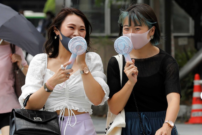 Two women wearing masks blow fans on their faces as they walk