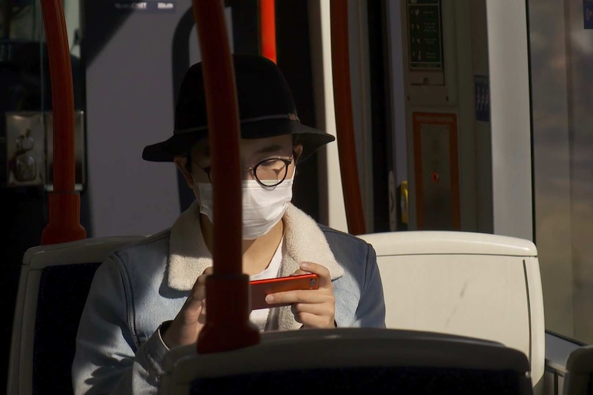 A man wearing a face mask sits on a tram using his mobile telephone.