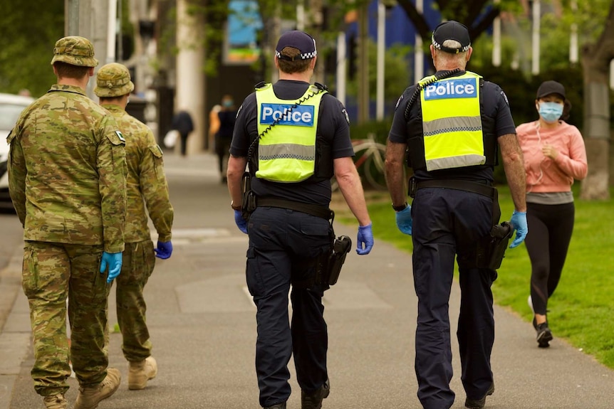 Two ADF officers and two police can be seen from behind patrolling a park as a woman jogs past wearing a face mask.