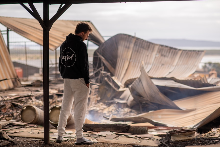 A man stands with his back to the camera, looking at rubble and twisted fencing