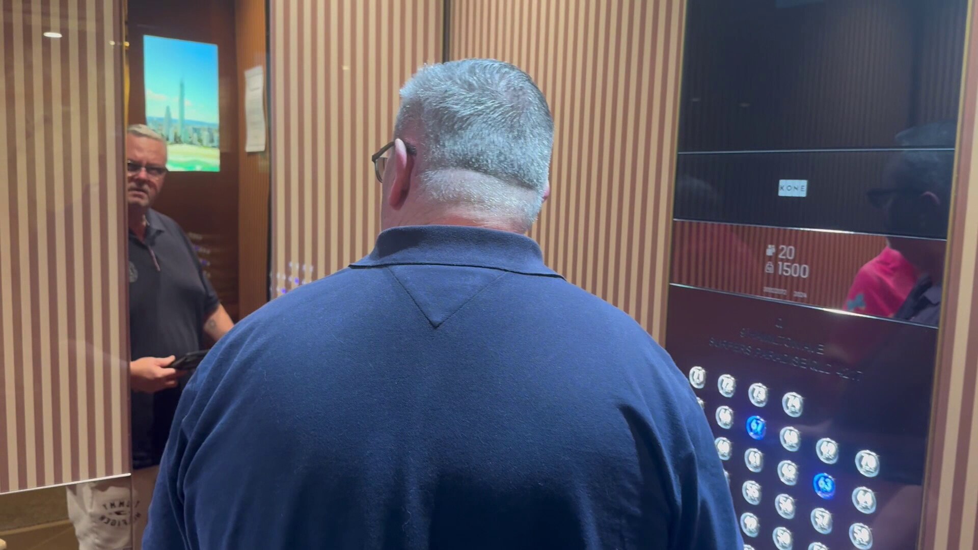 Un hombre vestido con una camisa azul oscuro está en un ascensor, de espaldas a la cámara. Un espejo refleja su rostro y muestra que lleva gafas.