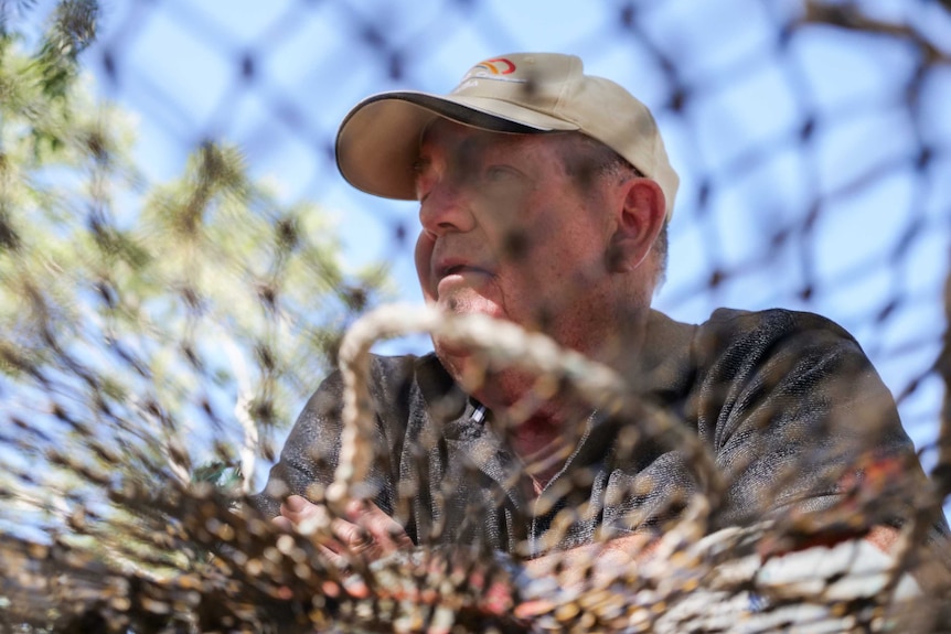 A man wearing a cap leans on a crabbing pot, the photo is taken through the pot, which is out of focus.