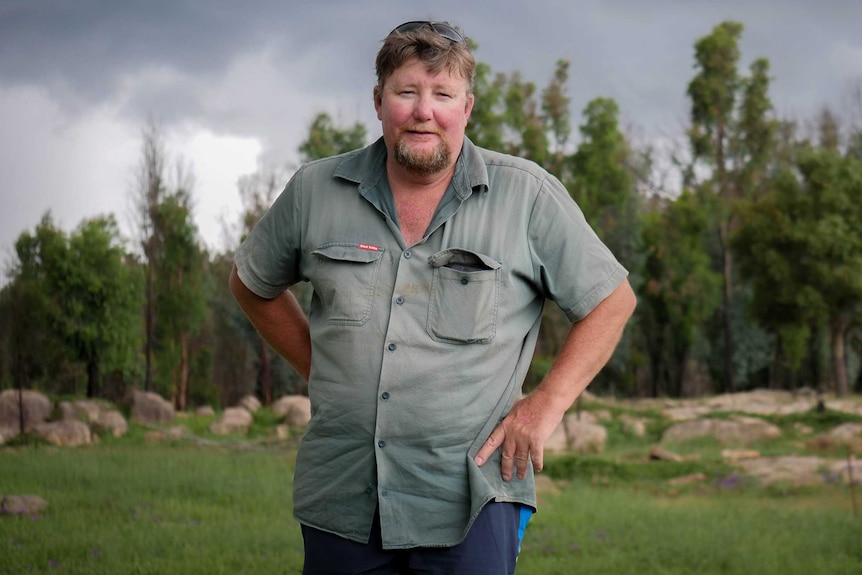A middle-aged white male farmer stands in a green paddock with a storm brewing behind him.