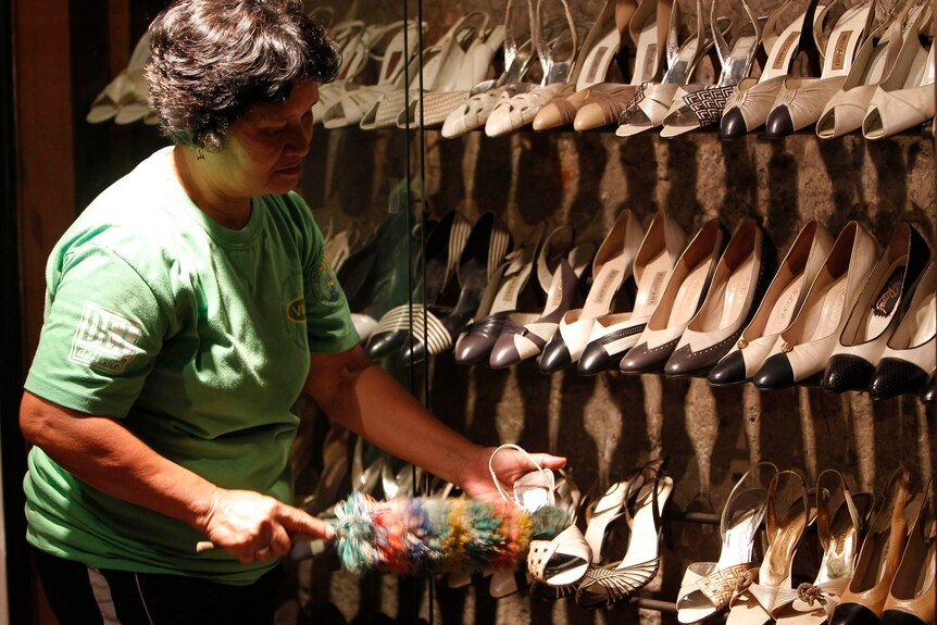 A woman dusts a display containing dozens of pairs of shoes.