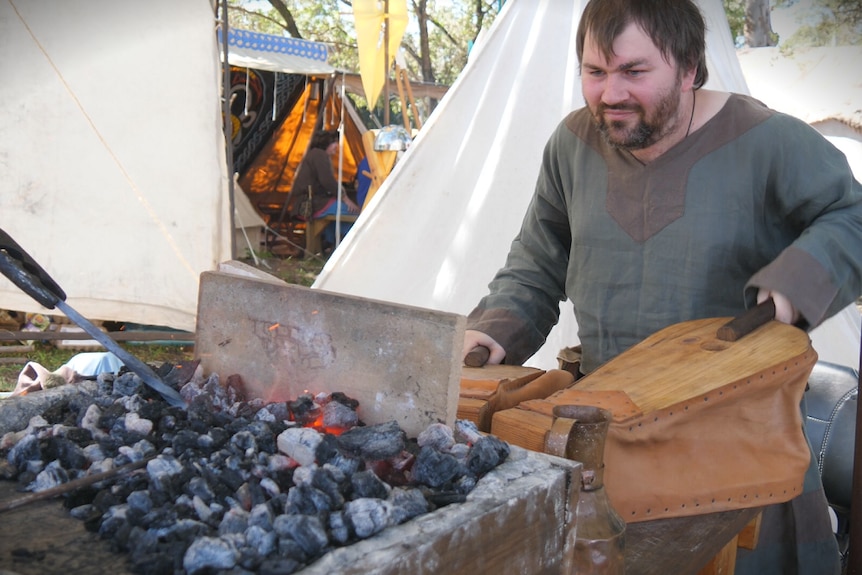 A man in a green shirt is holding leather and wood pumps that push air into hot coals.