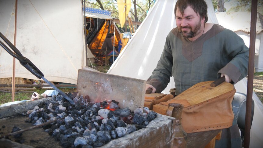 A man in a green shirt is holding leather and wood pumps that push air into hot coals.