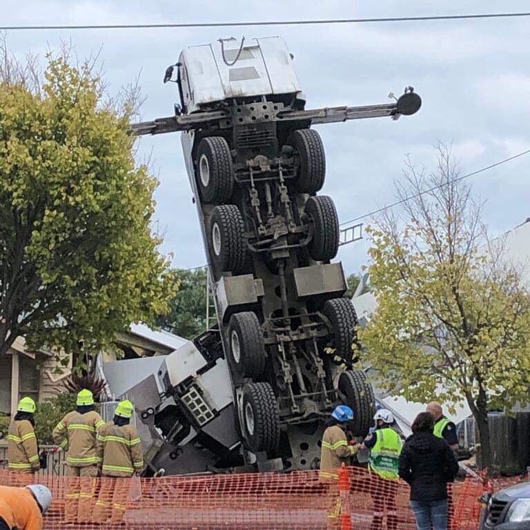 A crane stands upright in a residential street while firefighters gather around it.