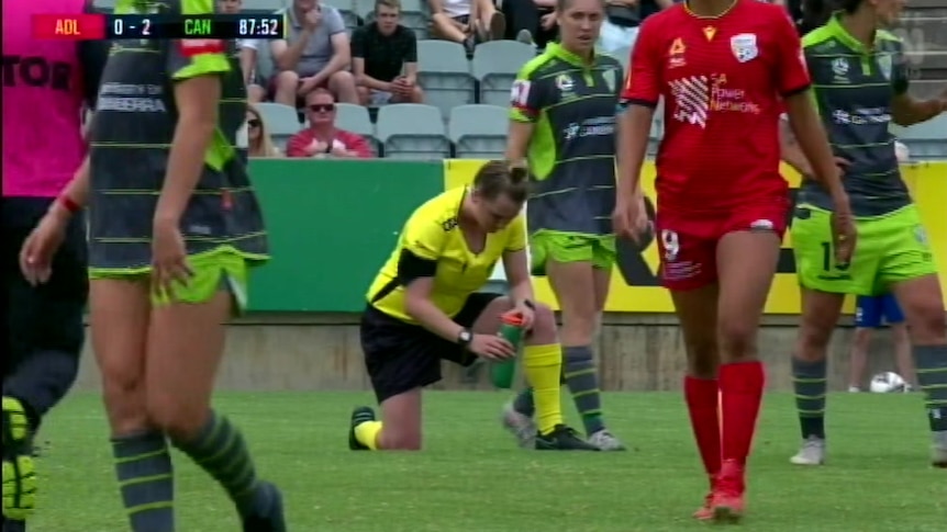 A woman in bright yellow clothes doubles over, unwell, on a soccer pitch.