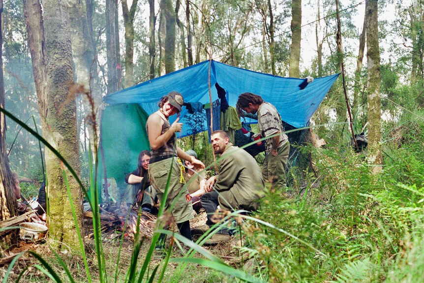 a group of young people in alternative clothing sit under a tarp cooking in the forest. 