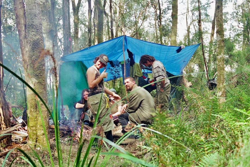 a group of young people in alternative clothing sit under a tarp cooking in the forest. 