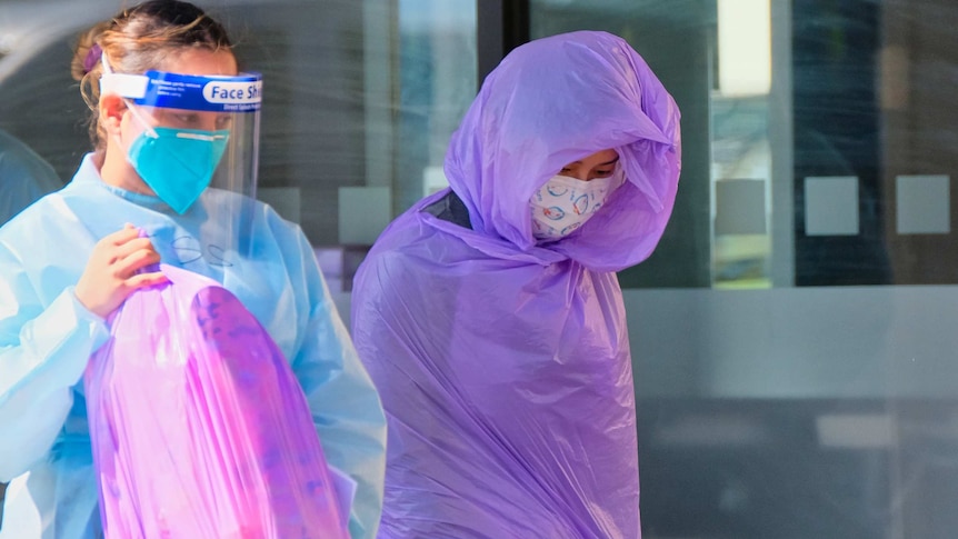 A woman swaddled in plastic and another woman in full PPF holding a full plastic bag.
