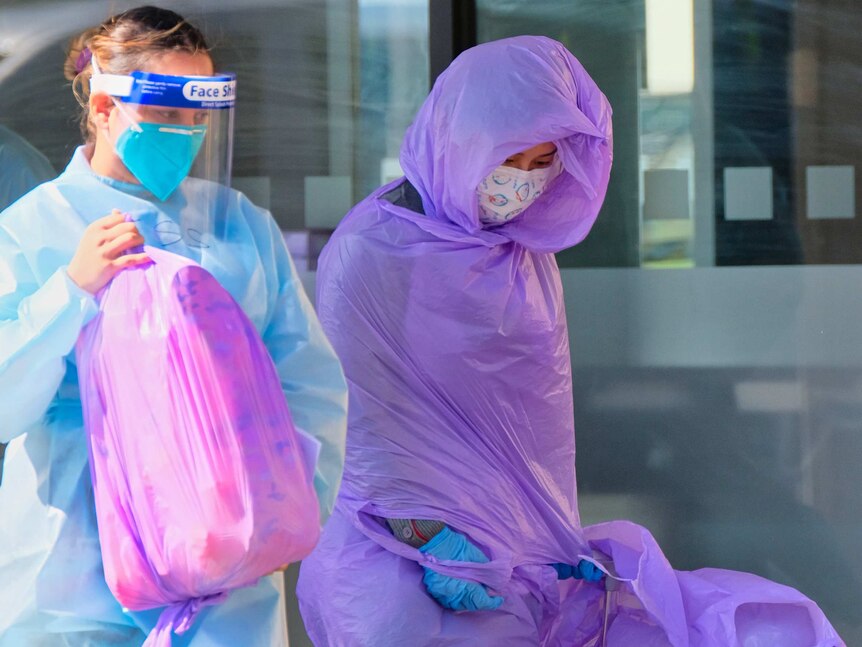 A woman swaddled in plastic and another woman in full PPF holding a full plastic bag.