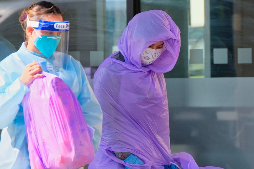 A woman swaddled in plastic and another woman in full PPF holding a full plastic bag.