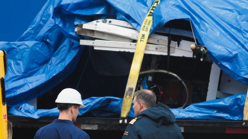 A closeup of debris from the Titan submersible pictured under a blue tarp, tied down with yellow straps.