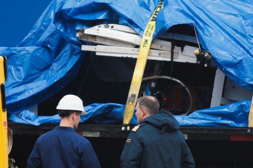 A closeup of debris from the Titan submersible pictured under a blue tarp, tied down with yellow straps.